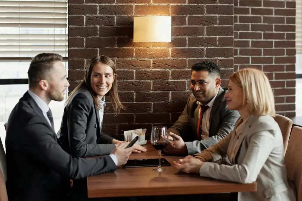 Four business professionals sitting around a table, smiling and chatting in a lively manner while enjoying drinks, conveying a warm and friendly atmosphere during a business meeting. They know about the French Etiquette for business.