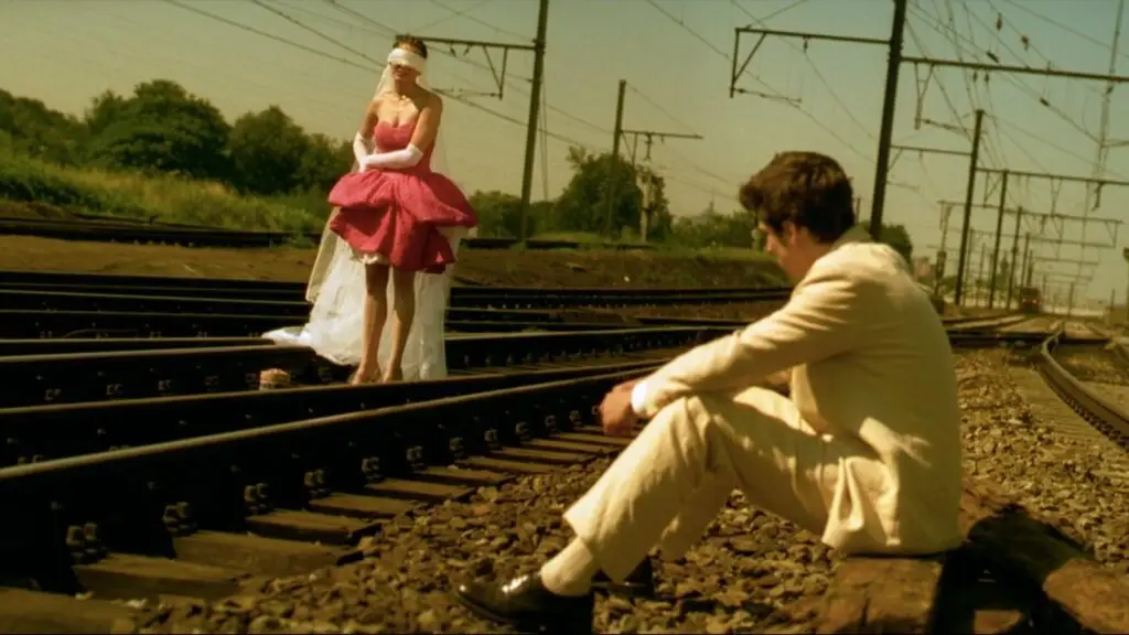 A dramatic scene from Jeux d’enfants (Love Me If You Dare) (2003). A woman in a beautiful dress stands on train tracks with her eyes covered, embodying the thrill and recklessness of their lifelong game of dares. A man sits nearby, observing her with a mix of amusement and tension, highlighting their intense, playful bond.