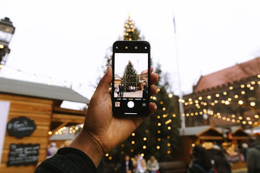 A hand holding a smartphone captures a holiday scene with a Christmas tree and market lights.