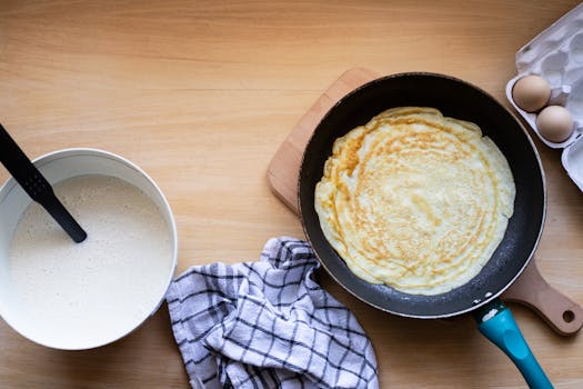 Top view of a pancake cooking in a pan with batter and eggs nearby.